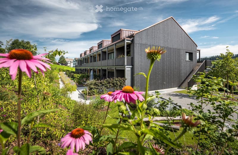 Modern architectural building with black exterior, multiple levels, balconies, surrounded by landscaped garden with vibrant flowers.