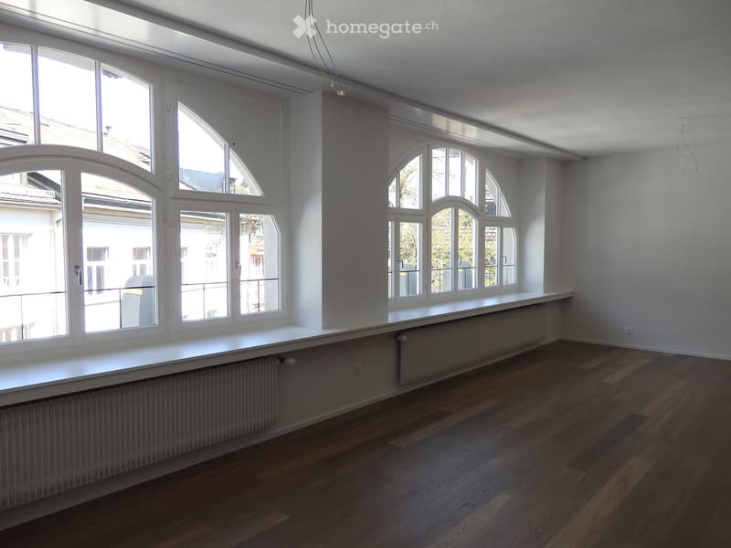 Empty room with wooden flooring, white walls, large windows, and radiators.