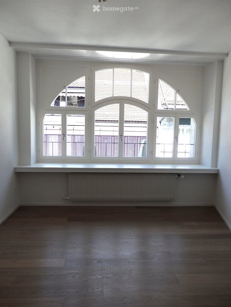 Empty room with wooden flooring, white walls, large windows, and a radiator below the windows.