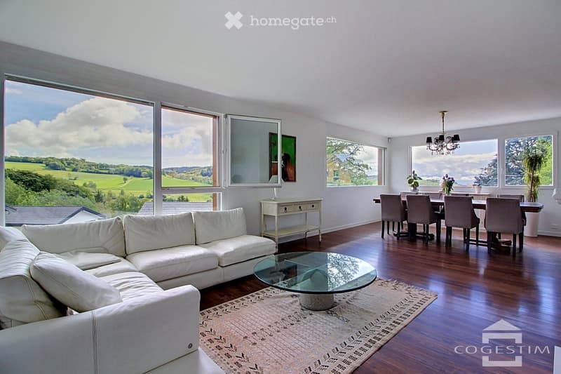 Bright living and dining area with large windows offering scenic views, featuring a white sectional sofa, glass coffee table, dining table with chairs, and a chandelier.