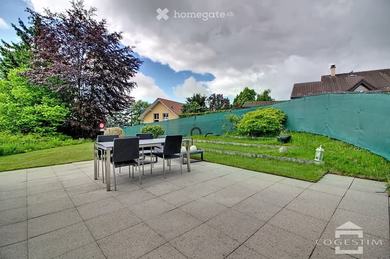 Garden with a patio area featuring a table and chairs, surrounded by greenery and a fence.
