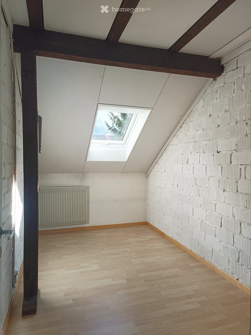 Empty attic room with wooden flooring, exposed wooden beams, white brick wall, skylight window, and radiator.