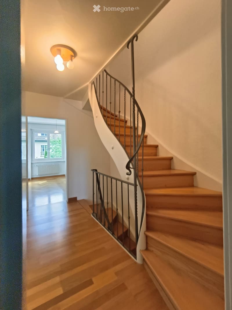 Spiral staircase with wooden steps and metal railing, leading to a bright room with large windows and wooden flooring.