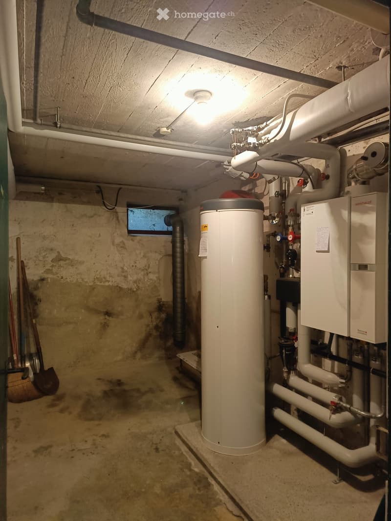 Utility room with exposed concrete walls, a water heater, pipes, and a small window. Tools are stored on the left side.