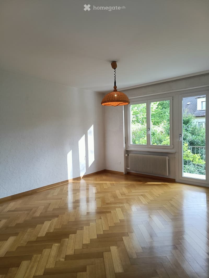 Empty room with wooden herringbone flooring, white walls, large windows, and a hanging pendant light fixture.