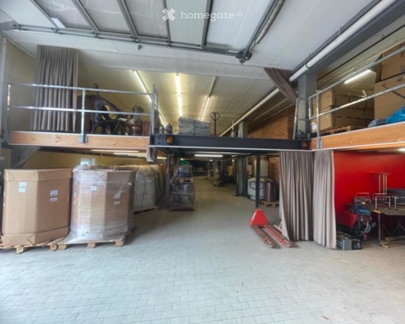 Large industrial-style warehouse space with high ceilings, exposed metal beams, and a mezzanine level. The floor is tiled, and the walls are painted in neutral tones. The space is mostly empty, with only a few stacked boxes and a small table visible.