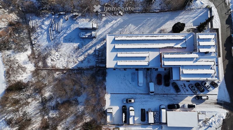 Aerial view of a large industrial building with a flat roof, surrounded by a parking lot with several vehicles and storage containers.