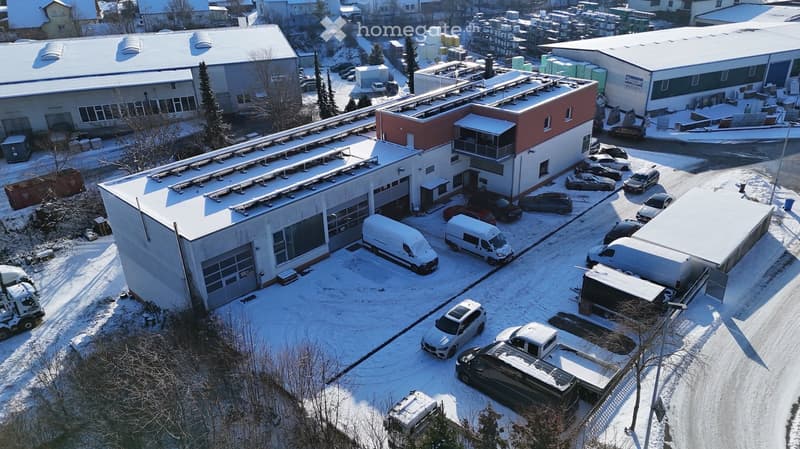 Aerial view of a large industrial building complex with multiple structures, snow-covered roofs, and a parking lot filled with vehicles.