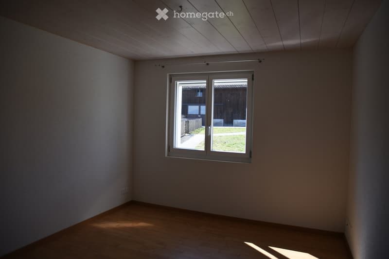 Empty room with wooden flooring, white walls, and a large window offering a view of a grassy area and wooden fence.