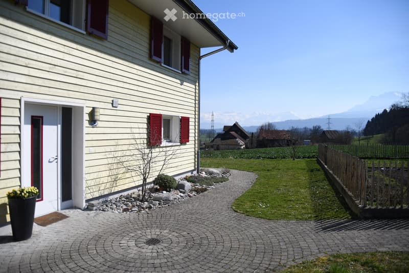 Front garden area with a paved pathway leading to the house entrance, small plants, a wooden fence, and a scenic mountain view in the background.