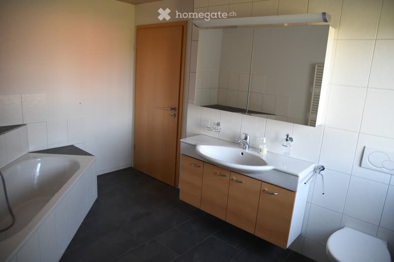 Modern bathroom with a bathtub, sink, mirror, and toilet. The sink has a wooden cabinet underneath, and the walls are tiled.