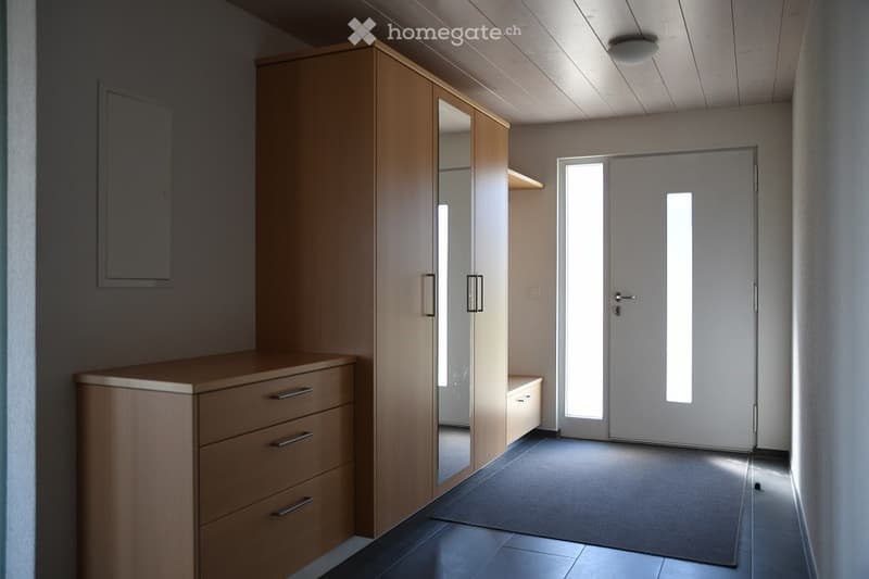 Empty room with wooden furniture including a tall wardrobe with mirrored doors, a chest of drawers, and a small bench. The room has a tiled floor and a large white door with glass panels.