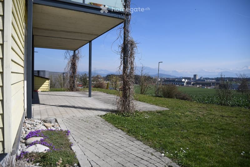A paved pathway leads to a covered balcony area with a view of a grassy lawn, distant fields, and mountains. Small purple flowers are planted along the pathway.