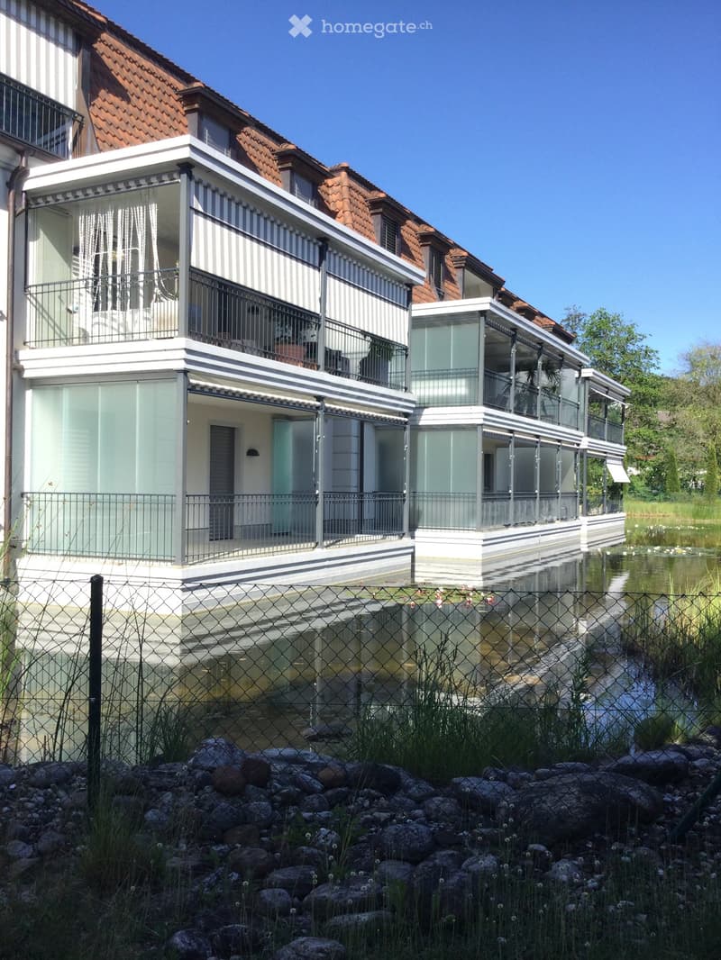 Modern two-story house with white and grey exterior, large glass balconies, and a red-tiled roof, situated near a water body with a fence and rocks in the foreground.