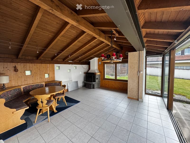 Furnished Room. Cozy living room with wooden beams, a round wooden table with chairs, a wooden bench, a fireplace, and large windows overlooking a green area.