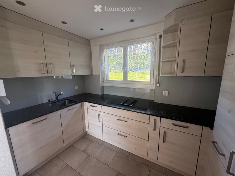 Kitchen. Modern kitchen with light wood cabinets, black countertops, a sink, and a stove. The space features a window with a decorative curtain and recessed ceiling lighting.