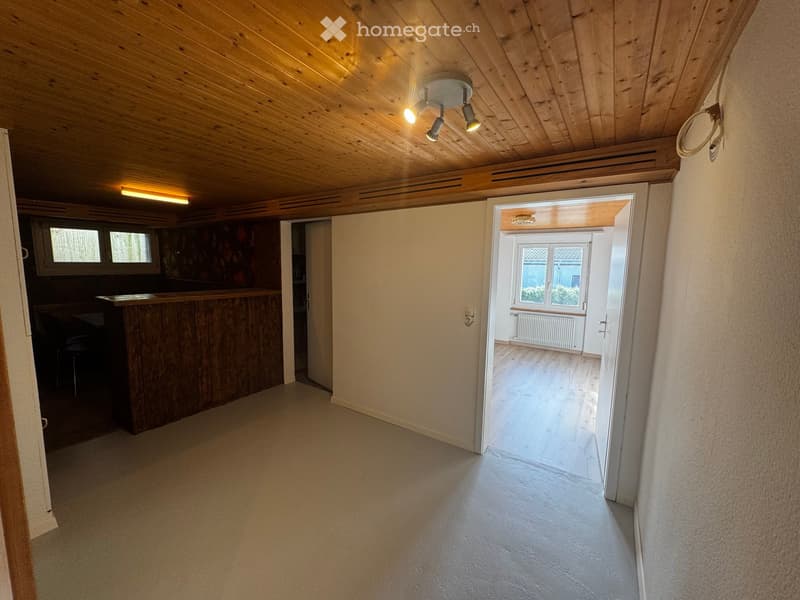 Corridor / Hallway. Empty hallway with wooden ceiling, light-colored walls, and a bar area visible to the left. The hallway leads to another room with a window and radiator.