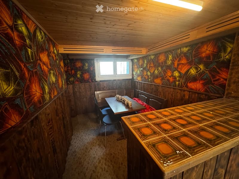 Furnished Room. A cozy wooden-paneled room with a table and chairs, colorful patterned wallpaper, and a window letting in natural light.