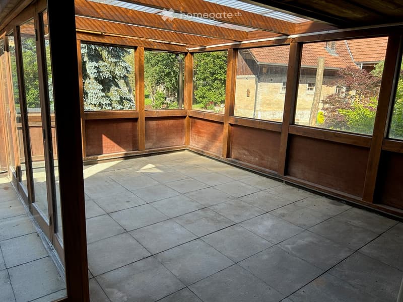 Empty wooden-framed balcony with tiled floor, large windows, and open sides, overlooking a garden and neighboring houses.