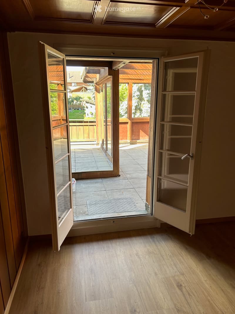 Empty room with wooden flooring and large open doors leading to a patio area with tiled flooring and wooden decking.