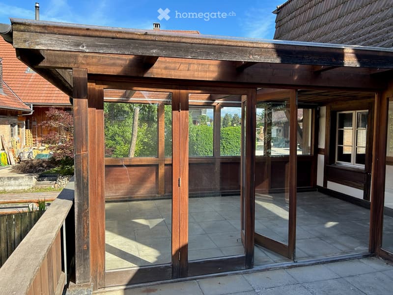 Wooden balcony with glass panels, tiled floor, and wooden posts, attached to a house with a tiled roof.