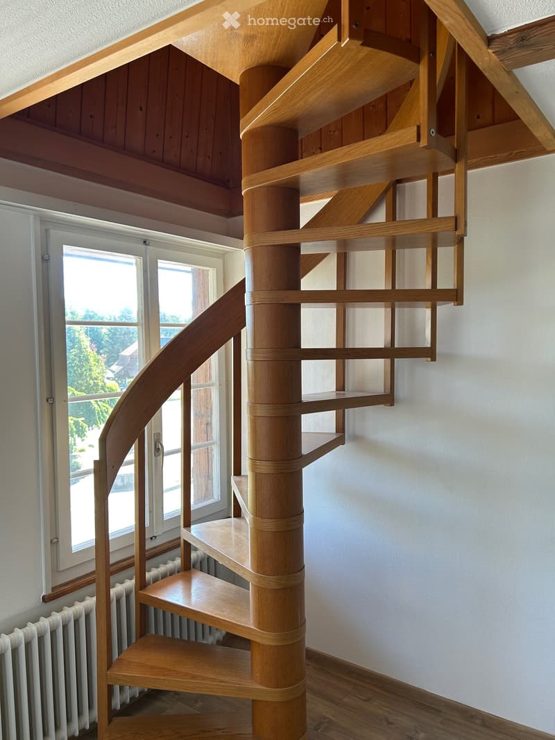 Spiral staircase with wooden steps and a central wooden column, leading to an upper level. The staircase is set against a white wall with large windows providing natural light.