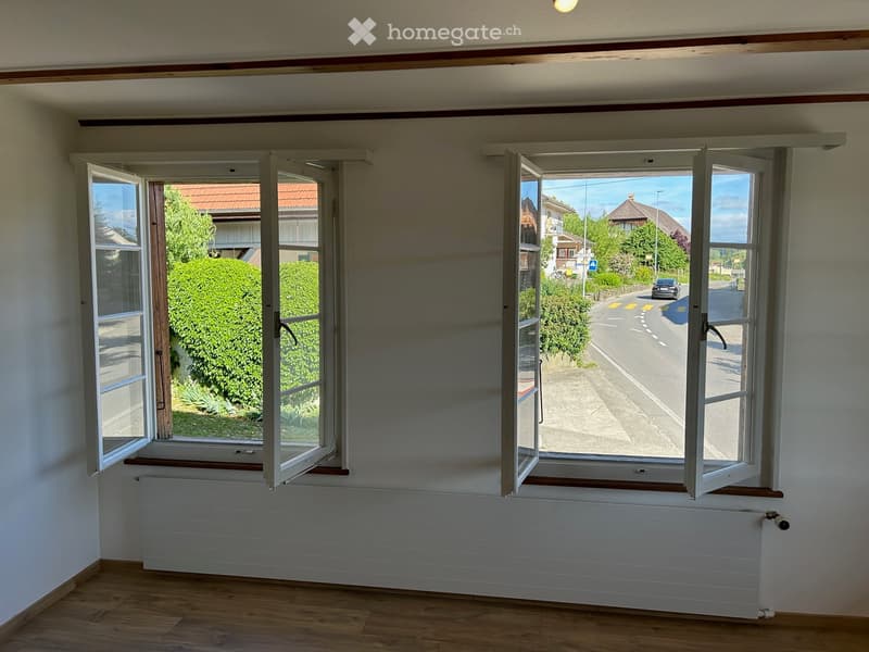 Empty room with wooden flooring, white walls, large windows with open shutters, radiator, and a view of the street and greenery.