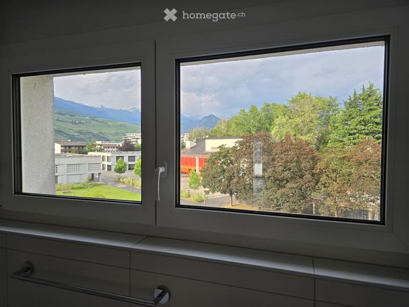 Bathroom with large windows offering a scenic view of mountains, trees, and buildings. The windows are framed in white and have a modern design.