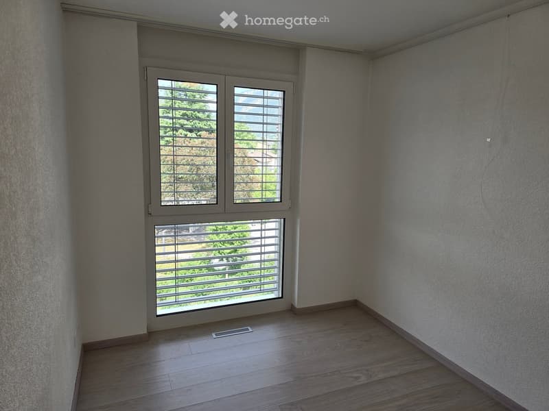 Empty room with wooden flooring, light grey walls, and large windows with blinds, offering a view of greenery outside.