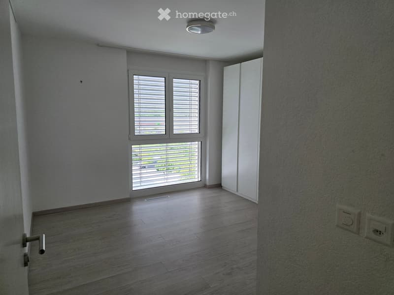 Empty room with wooden flooring, white walls, large window with blinds, and a closed white door.