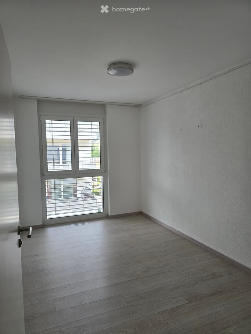 Empty room with light grey walls, wooden flooring, and a large window with blinds. The room has a modern ceiling light fixture and a door with a handle visible on the left side.