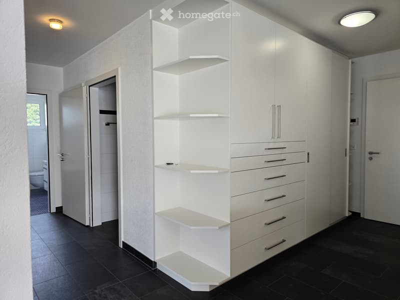 Modern hallway with white built-in storage units, open shelving, and recessed lighting. The hallway features dark tiled flooring and leads to a bathroom with a visible toilet and bathtub.