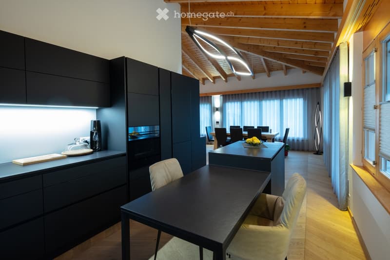 Modern kitchen and dining area with dark cabinets, a central island, and a dining table with chairs. The space features wooden beams on the ceiling and large windows with curtains.