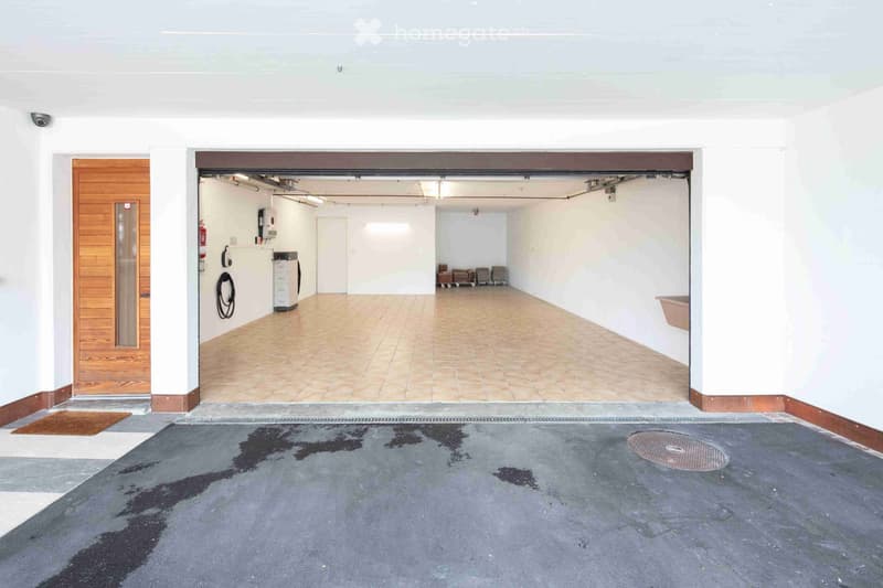 Empty garage with tiled floor, white walls, and a wooden door leading to another area. There are some stacked items in the background.