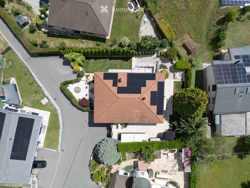 Aerial view of a residential area with multiple houses, solar panels on roofs, gardens, driveways, and parked cars.