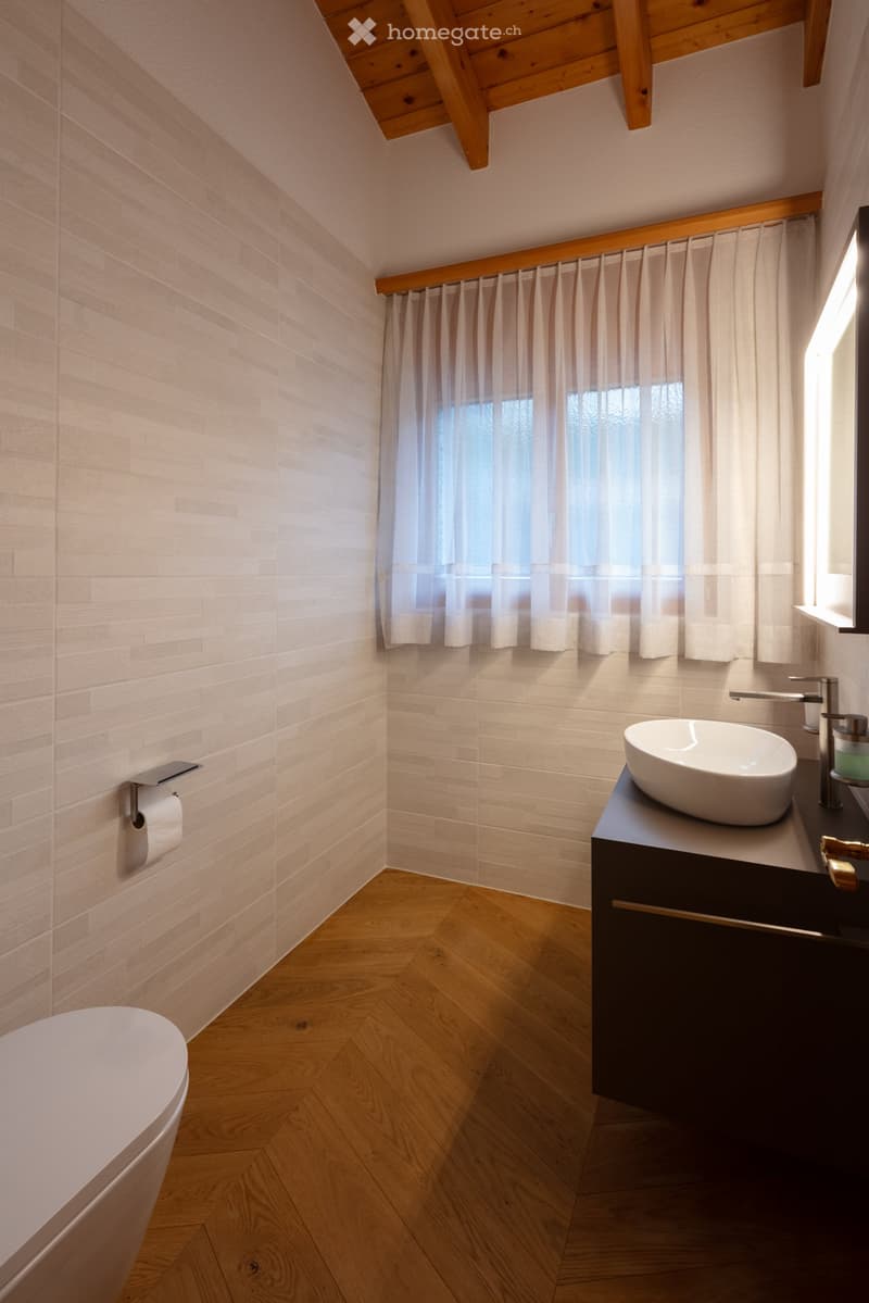 Modern bathroom with wooden herringbone flooring, light-colored tiled walls, a floating vanity with a sink, and a toilet. A window with sheer curtains allows natural light to enter.