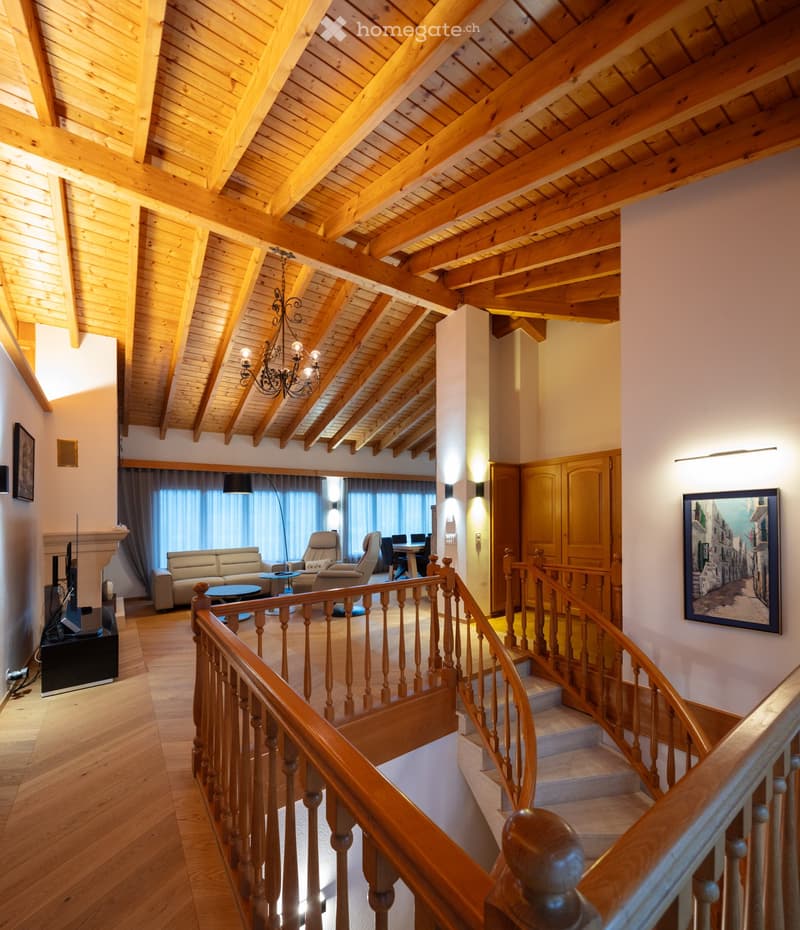 Spacious living area with wooden staircase, exposed wooden beams, chandelier, sofa, chairs, coffee table, TV, and artwork on walls.