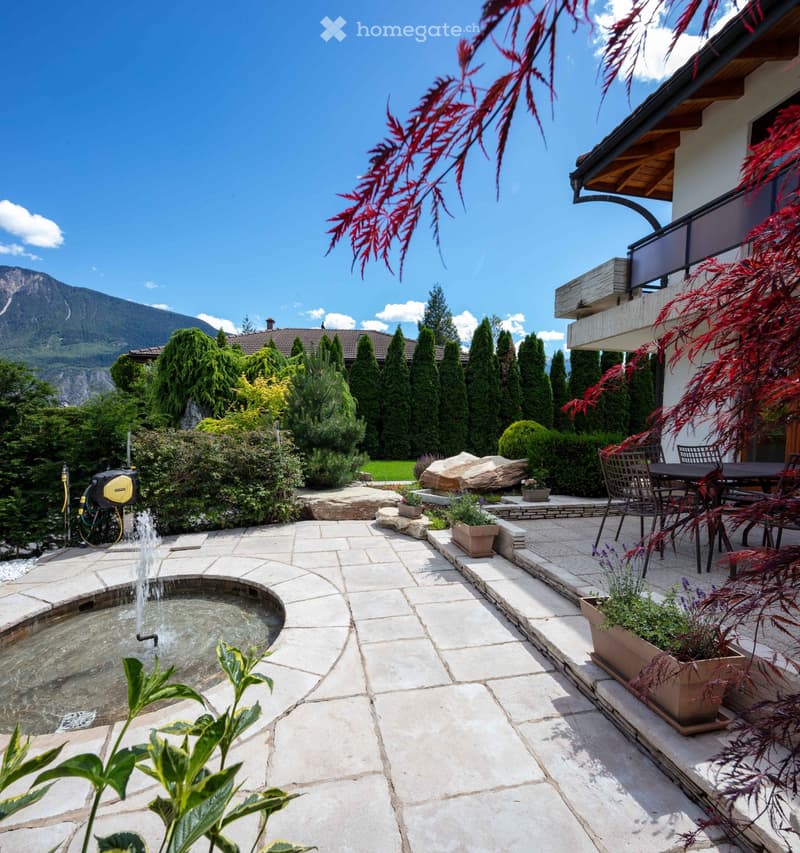 Well-maintained garden with a stone patio, fountain, potted plants, and outdoor furniture, overlooking a mountain view.