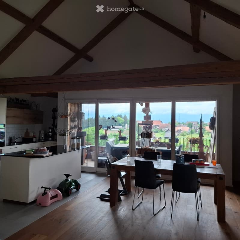 Furnished Room. Bright, open-plan room with wooden floors, a kitchen area, a dining table with chairs, and large windows offering a scenic view of the countryside.