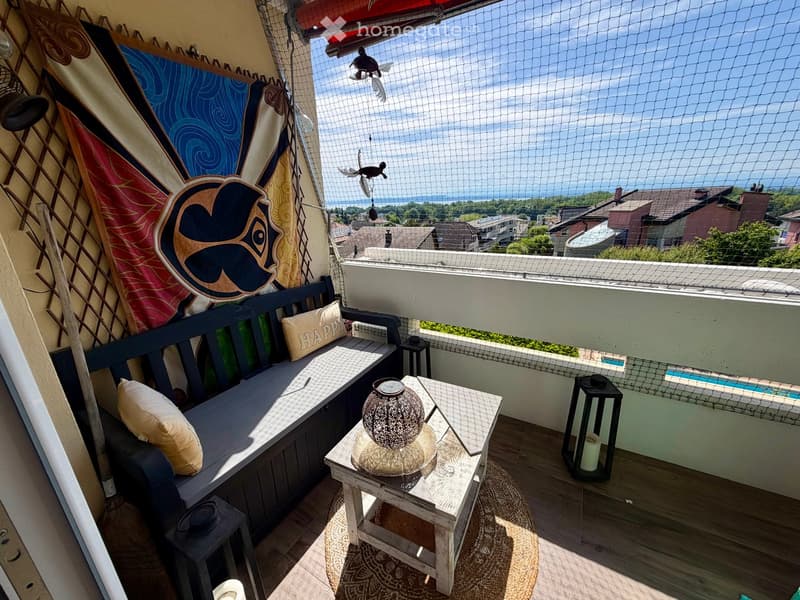 Balcony with a bench, small table, and decorative items, offering a scenic view of the ocean and surrounding area.
