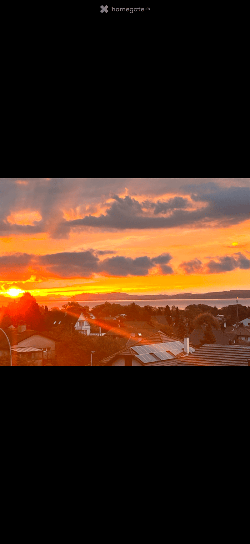 Residential neighborhood with houses, solar panels on roofs, and a scenic sunset view over a body of water.