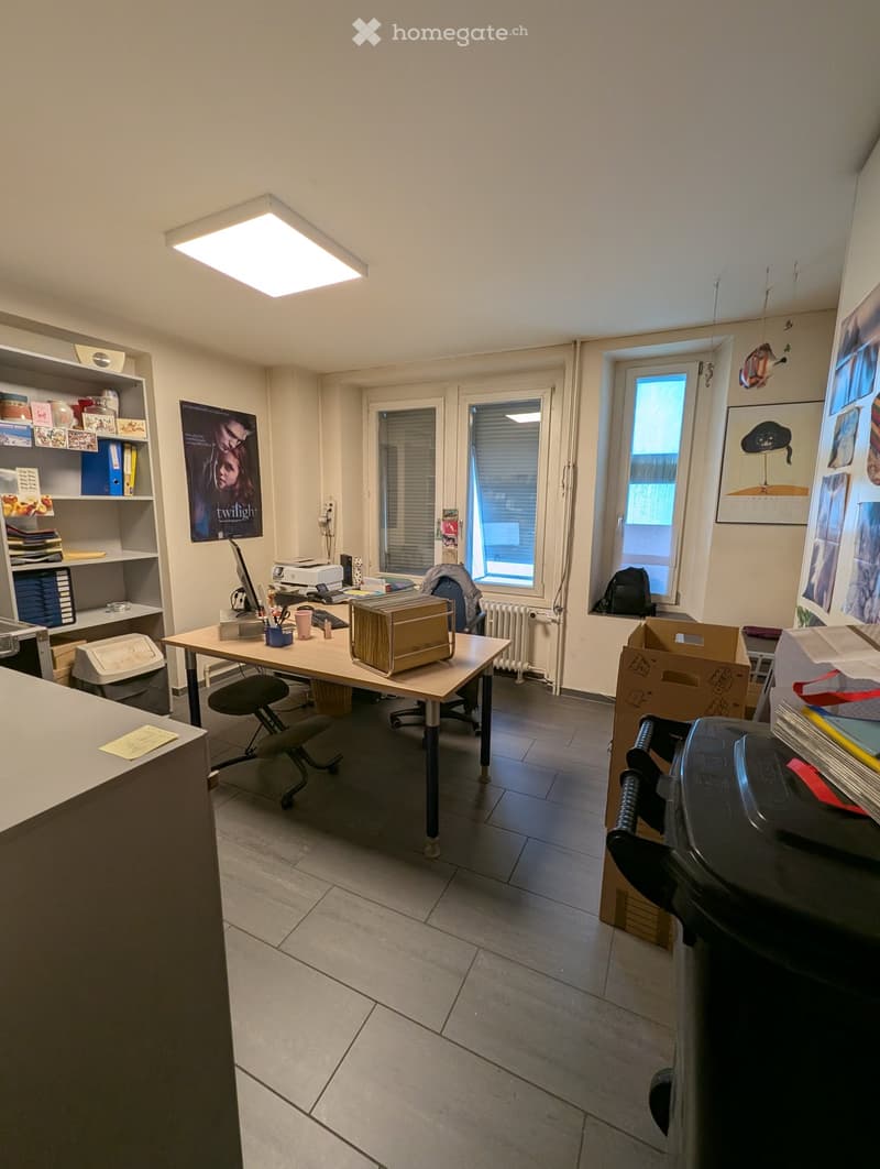 Office space with a desk, chair, shelves, printer, laptop, and various office supplies. The room has large windows, a radiator, and posters on the walls.