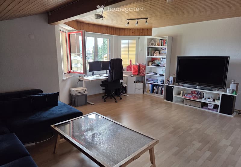 Modern living room with wooden floors, large windows, a dark blue sofa, a glass coffee table, a desk with a chair, a bookshelf, and a television on a stand.