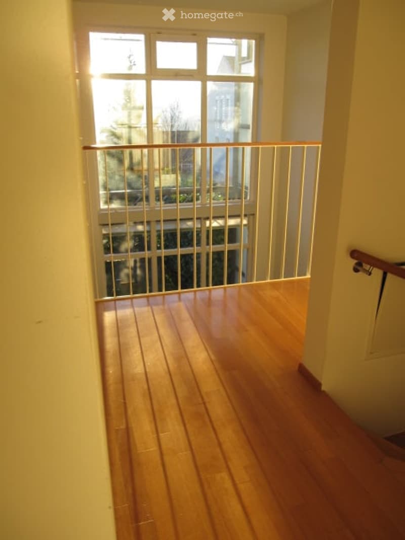 A narrow hallway with wooden flooring and a metal railing leading to a window with a view of the outside. The walls are painted a light color, and the space is well-lit with natural light coming through the window.