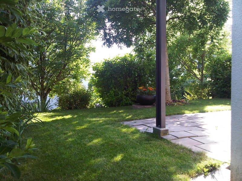 Well-maintained garden with a paved pathway, lush green grass, and various trees and shrubs. A large black planter is visible near the pathway.