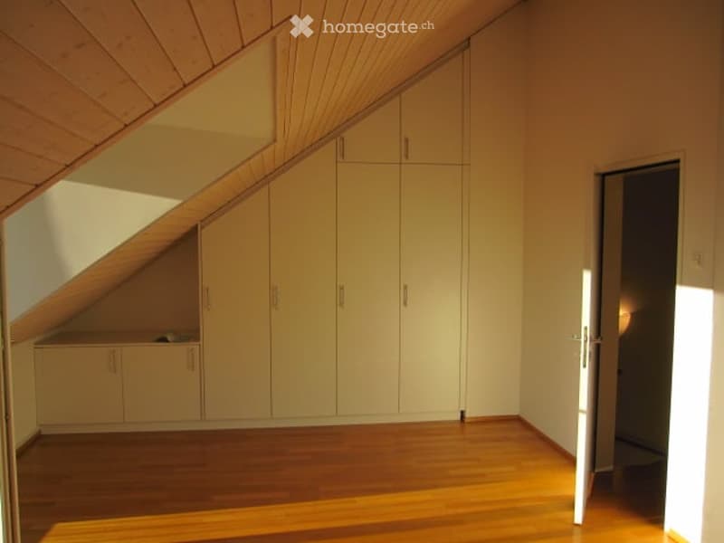 Empty attic room with wooden floor, white built-in wardrobes, and slanted wooden ceiling.
