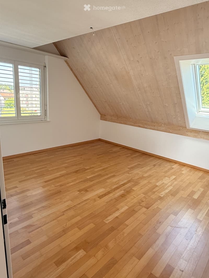Empty attic room with wooden flooring, white walls, and two windows with blinds. The ceiling has a sloped design with exposed wooden beams.