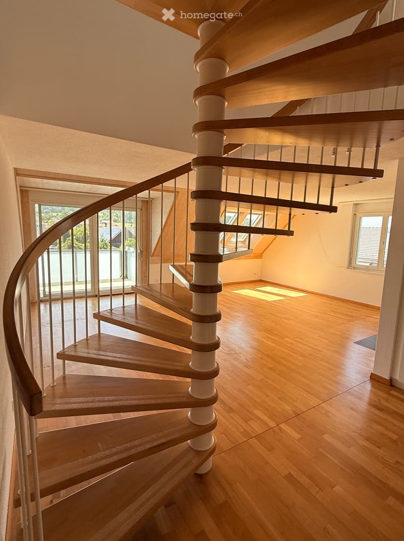 Spiral staircase with wooden steps and a white central column, leading to an open area with wooden flooring and large windows.