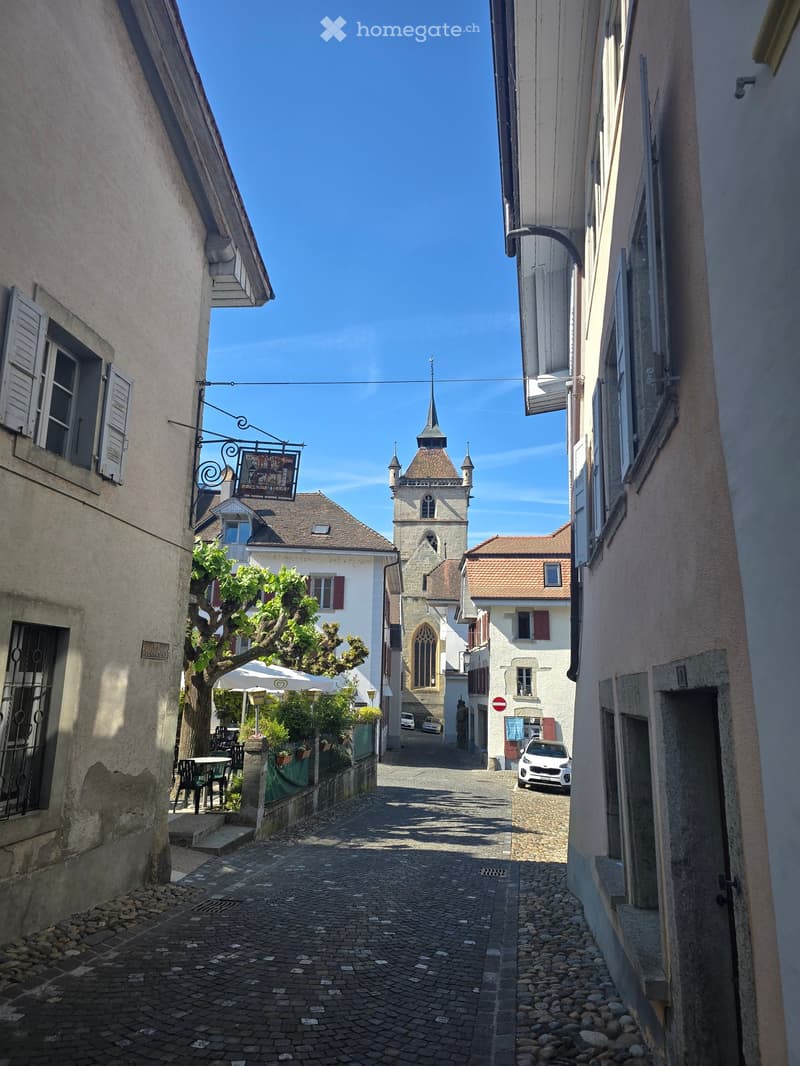 A narrow cobblestone street lined with old buildings, featuring a church tower in the background and a few parked cars.