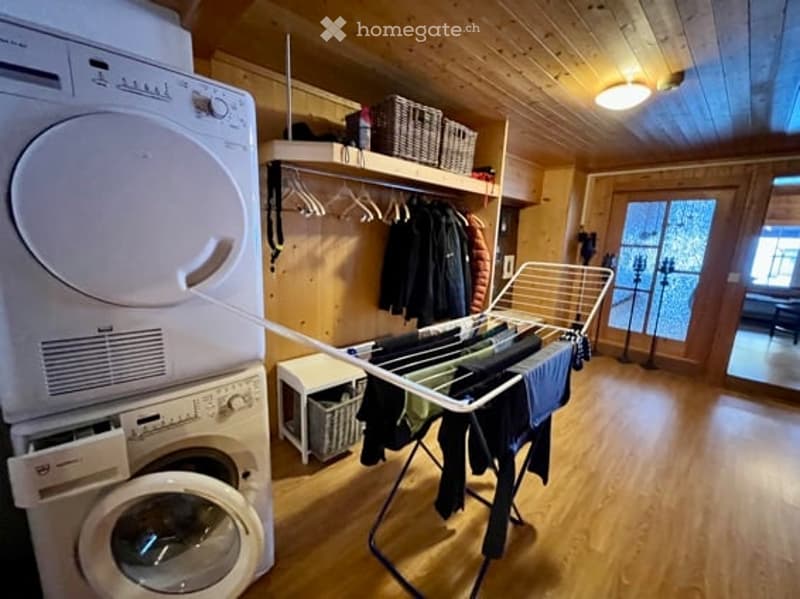 A cozy, wooden-walled room with a washing machine, drying rack, coat rack, and shelves holding baskets and other items.
